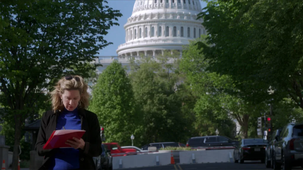The Capitol stands in the background as a woman wearing a blue shirt and black blazer writes on a red folder.