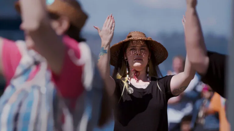 A woman wearing a woven hat and dark t-shirt stands outside, her arms raised in front of her. Other people surround her, making the same gesture.