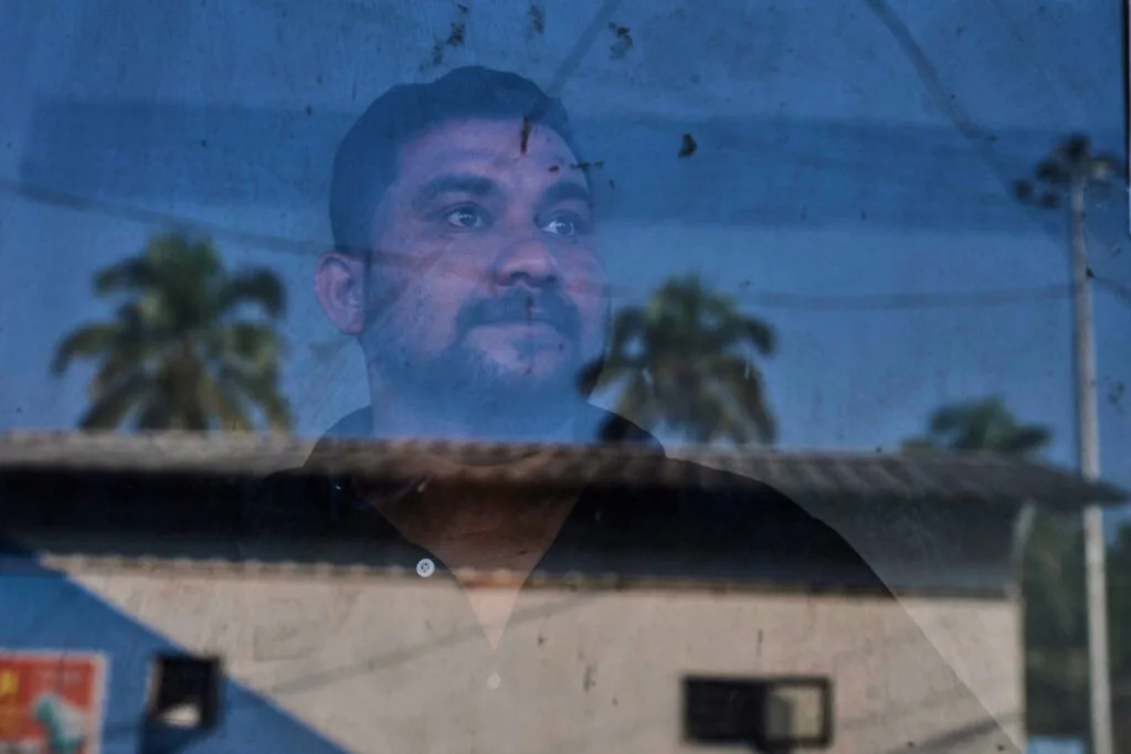 A man stands behind a pane of glass that reflects a building, palm trees and electric poles.