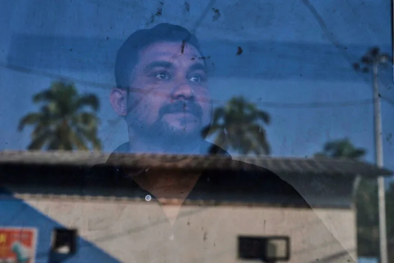 A man stands behind a pane of glass that reflects a building, palm trees and electric poles.