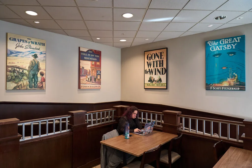 A woman sits in front of her laptop at a table with four chairs. Posters of vintage book covers hang on the store walls.