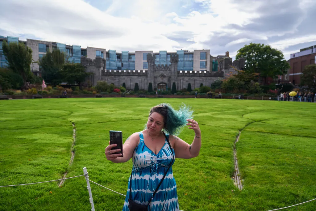 A woman stands on a grassy field with a castle in the background. She holds her phone while her hair blows in the wind.