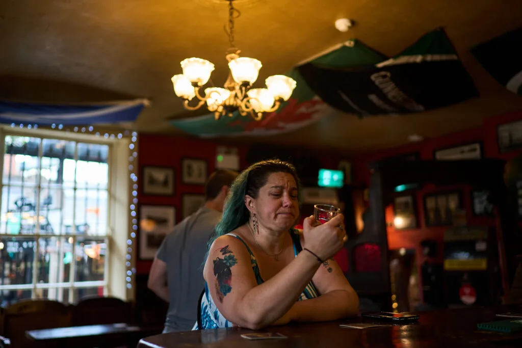 A woman sits in a bar holding a small glass and grimacing.
