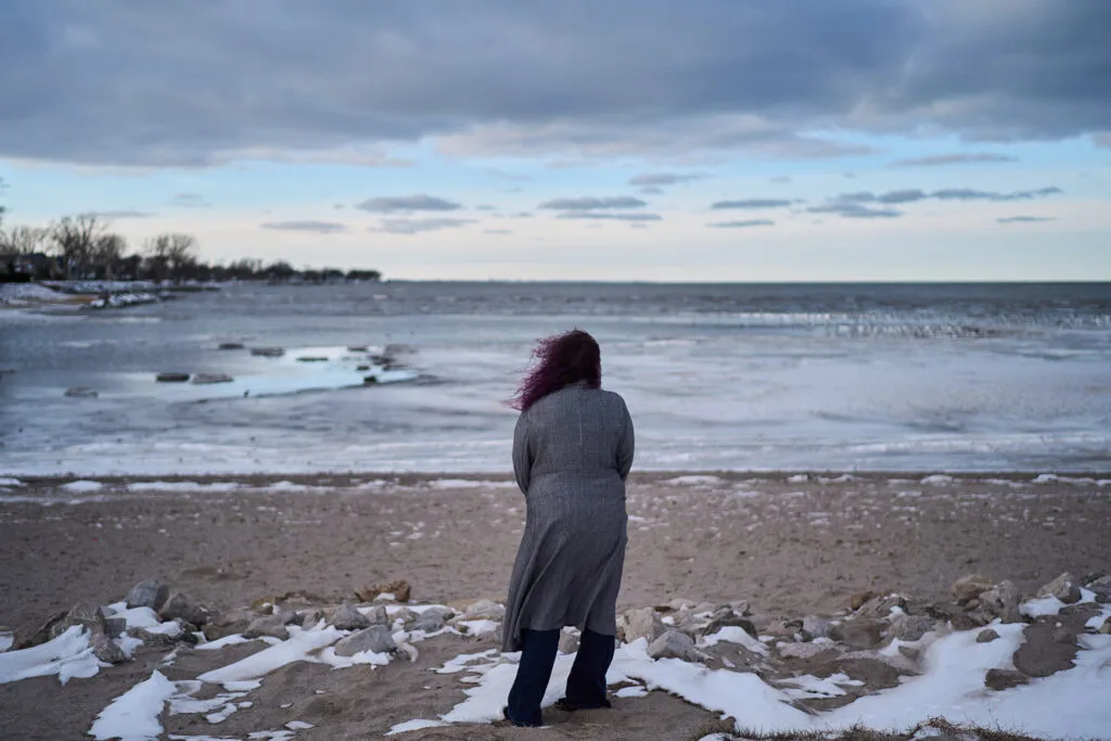 A woman with purple hair and a long winter coat is seen from behind, standing on a beach. It's winter, and there are patches of ice on the water and on land.