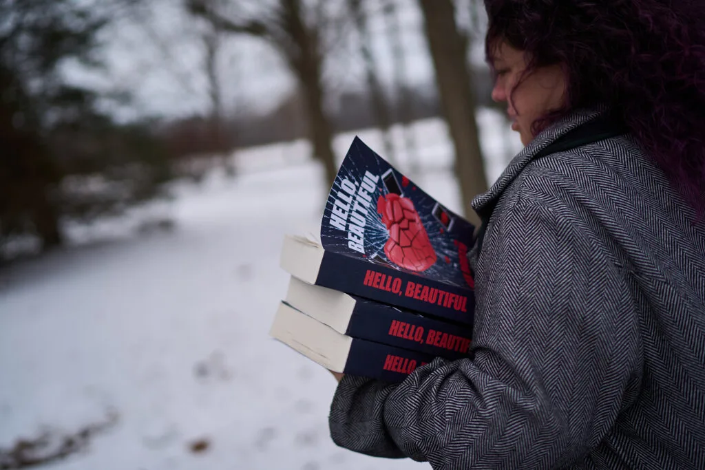 A woman wears a coat and holds books that have the title "Hello Beautiful" with a cover that has a shattering heart. She's standing outside in the snow.