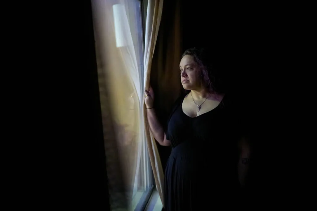 A woman stands at a window, looking out. The rest of the background is dark.
