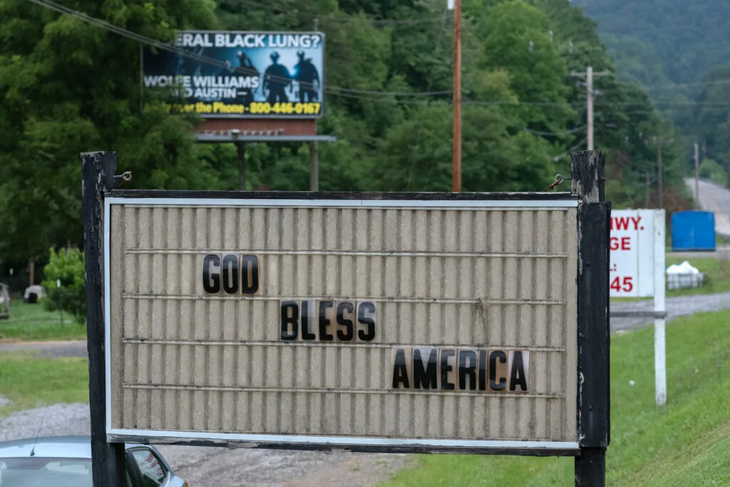 A weathered sign in the foreground reads "God bless America," and a billboard can be seen in the distance that has the words "black lung" on it.