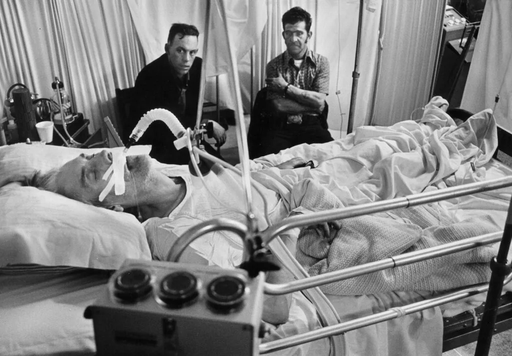 A black and white image shows a man lying in a hospital bed unconscious and intubated while two other men sit by his bedside.