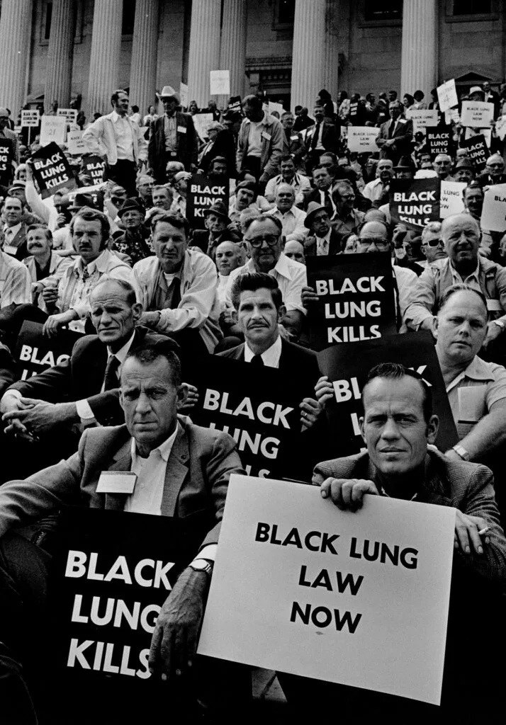 Dozens of men sit on the U.S. Capitol steps in a black and white image. Many hold signs that say "black lung kills," and one in the foreground holds a sign that says "black lung law now."