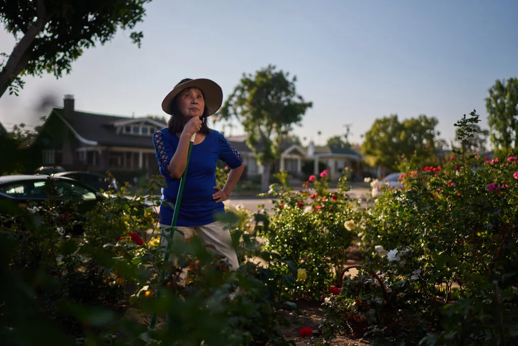 An older Asian woman wearing a blue shirt and hat holds a watering hose and stands in a garden.
