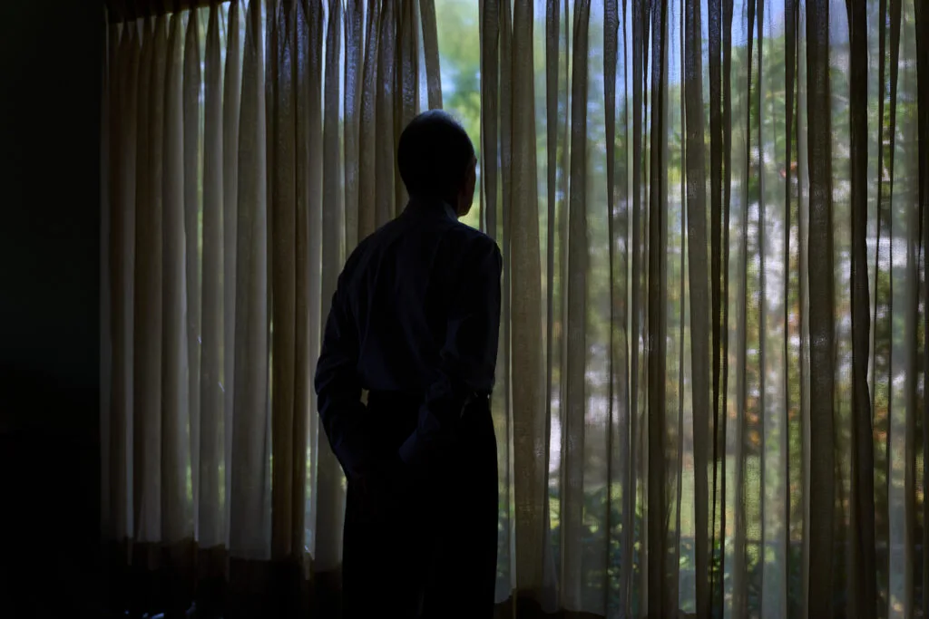 A person in a darkened room stands at a window looking out.