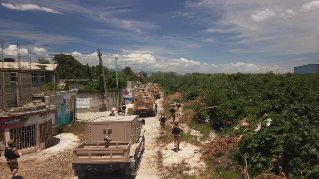 Armored military-style vehicles drive on a dusty road on a sunny day, with armed personnel walking next to them. On one side of the road are crumbling buildings and on the other is thick vegetation.