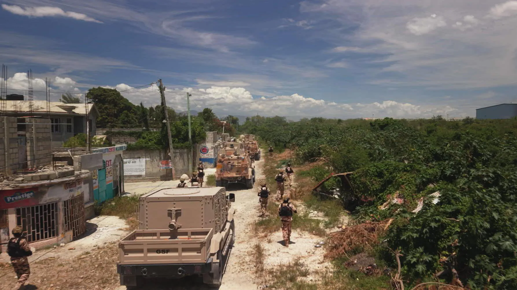 Armored military-style vehicles drive on a dusty road on a sunny day, with armed personnel walking next to them. On one side of the road are crumbling buildings and on the other is thick vegetation.