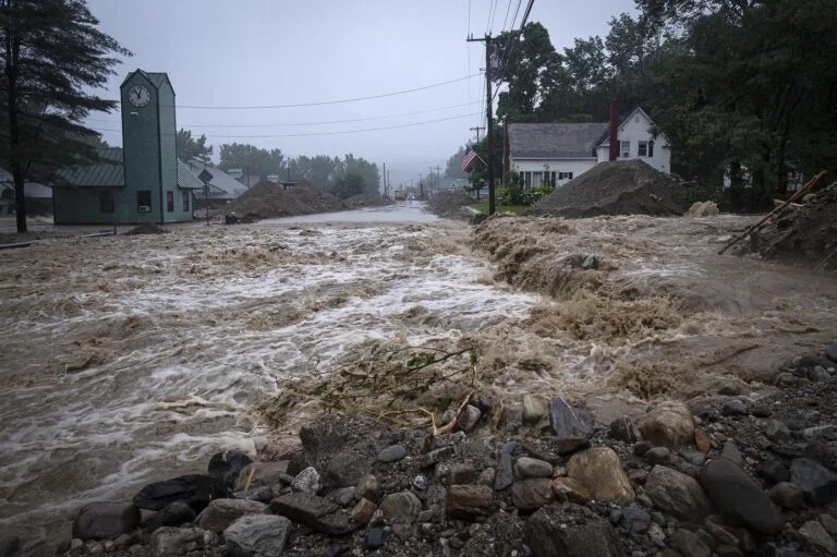 A street flooded with brown, churning water. Buildings in the distance have dirt piled around them.