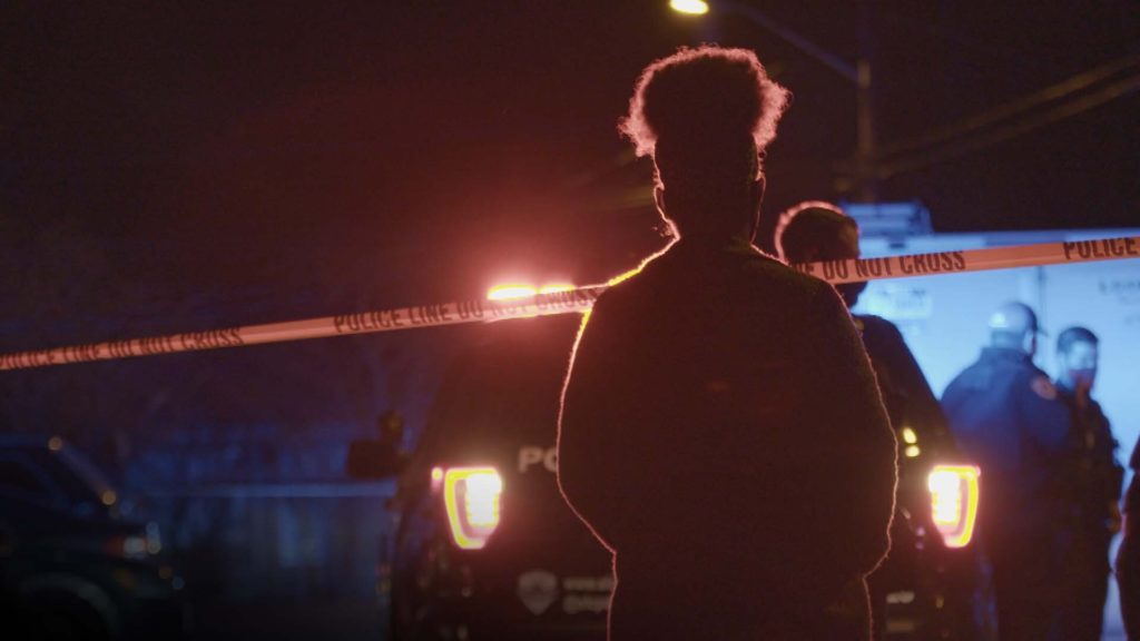 In a still image from the FRONTLINE and Salt Lake Tribune documentary "Shots Fired," on police shootings in Utah, a woman backlit by police headlights at night stands in front of police tape reading, "Police line do not cross."