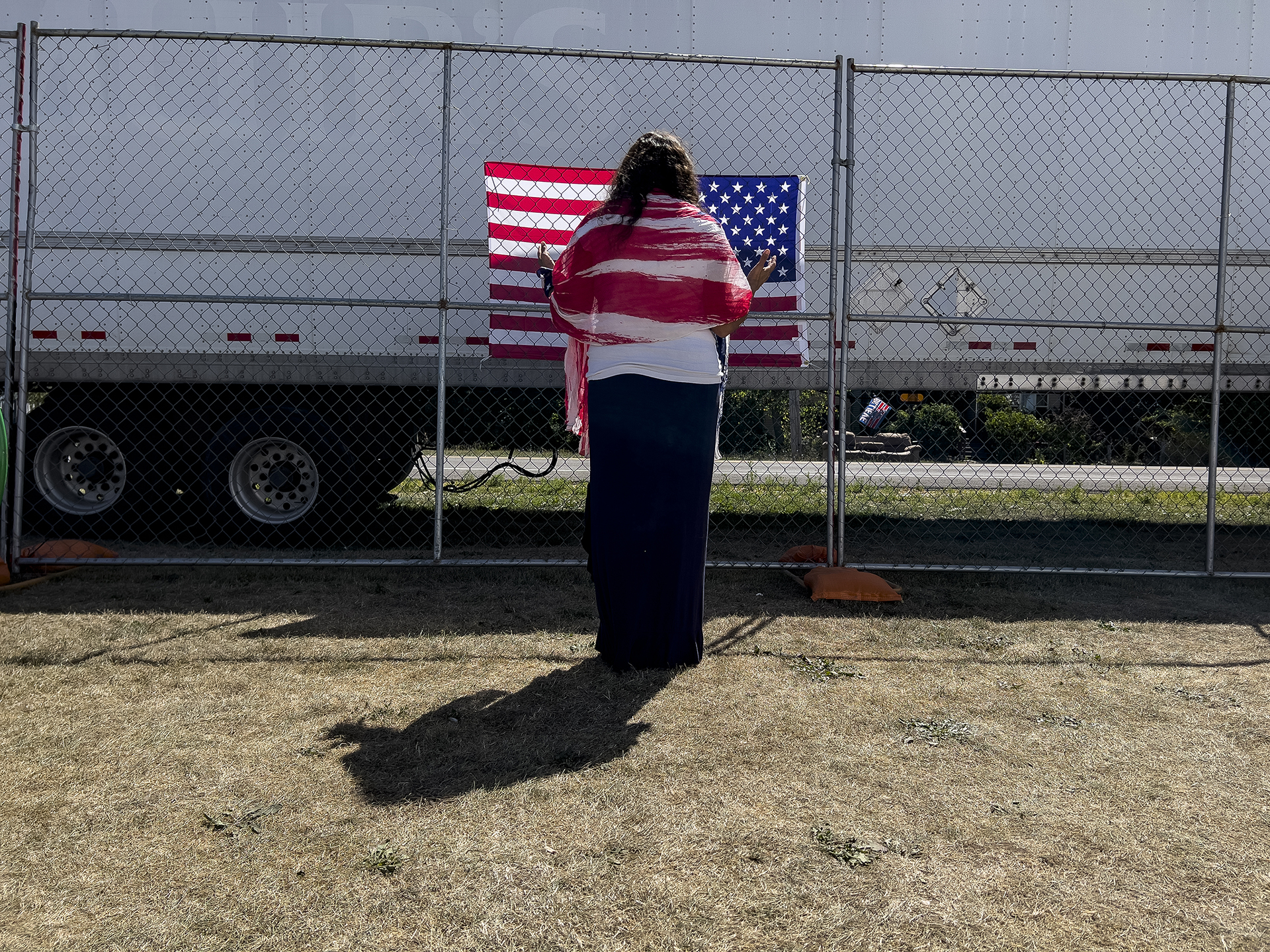 A woman prays as she faces an American flag on the perimeter fence at the ReAwaken America tour at Cornerstone Church in Batavia, N.Y., Saturday, Aug. 13, 2022. (AP Photo/Carolyn Kaster)