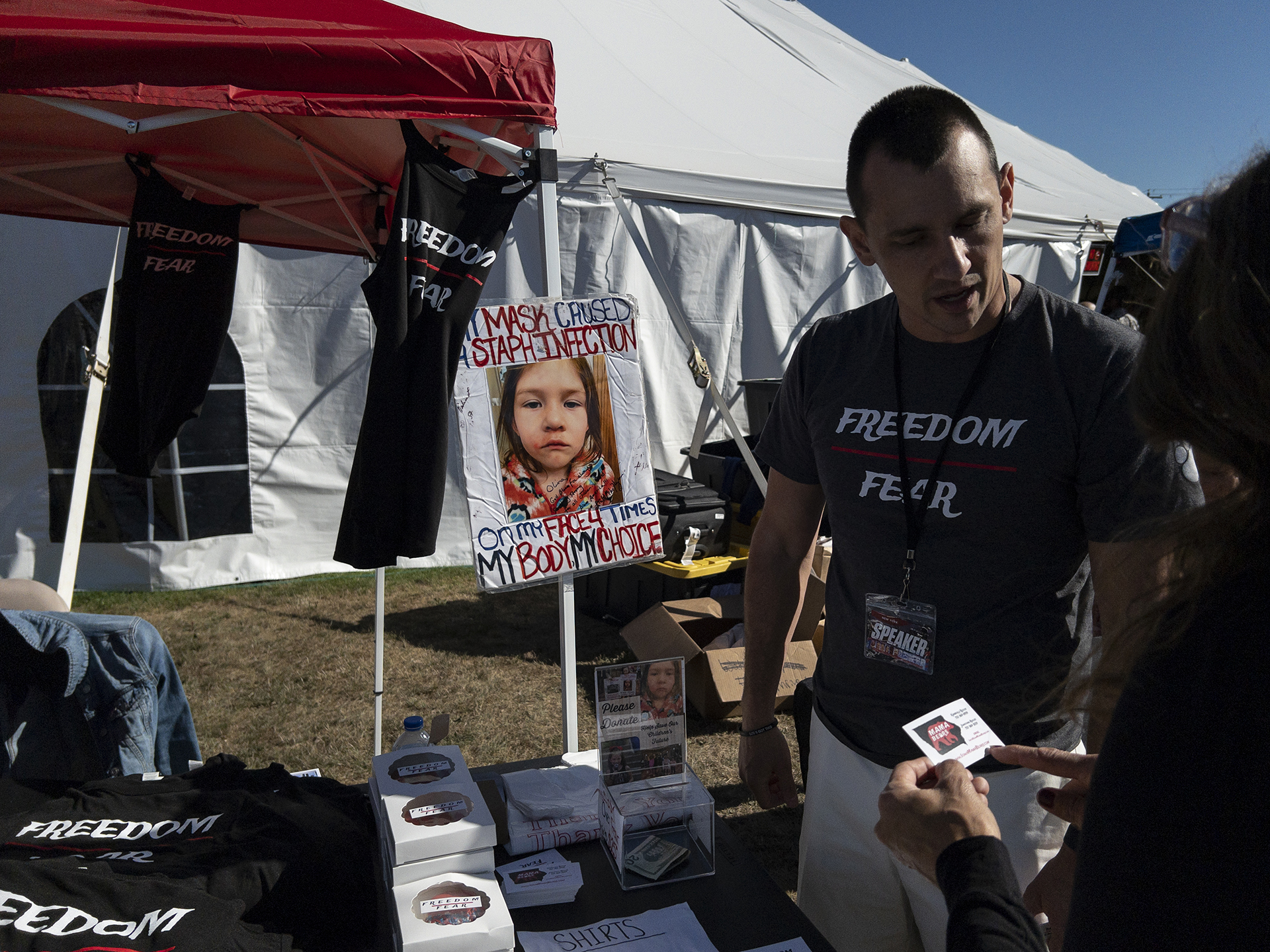 Jonathan Reicks talks at Iowa Mama Bears’ “Freedom Over Fear” merchandise table during the ReAwaken America tour at Cornerstone Church, in Batavia, N.Y., Friday, Aug. 12, 2022. A poster bearing a photo with Reicks’ kindergarten-aged daughter is displayed at their booth. (AP Photo/Carolyn Kaster)