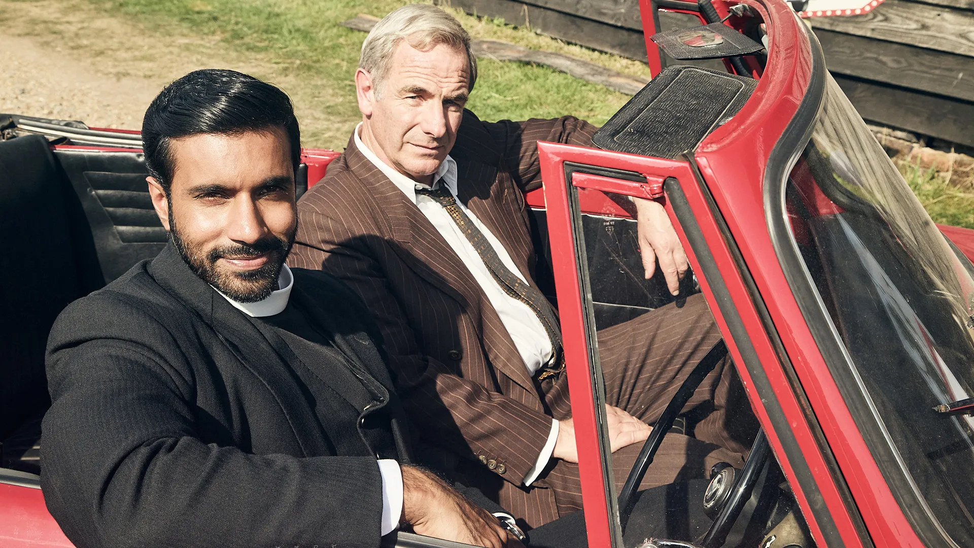 Color photo of actor Rishi Nair in costume as Rev Alphy Kottaram and actor Robson Green in costume as DI Geordie Keating in a scene from Grantchester Season 11.