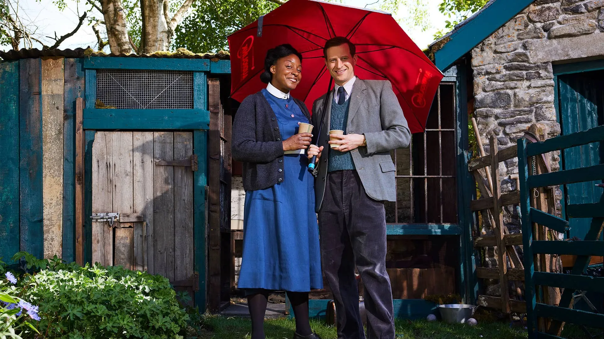Elexi Walker (Sister Rose) and Nicholas Ralph stand in costume under a red umbrella to protect them from the sun and heat, smiling at the camera.