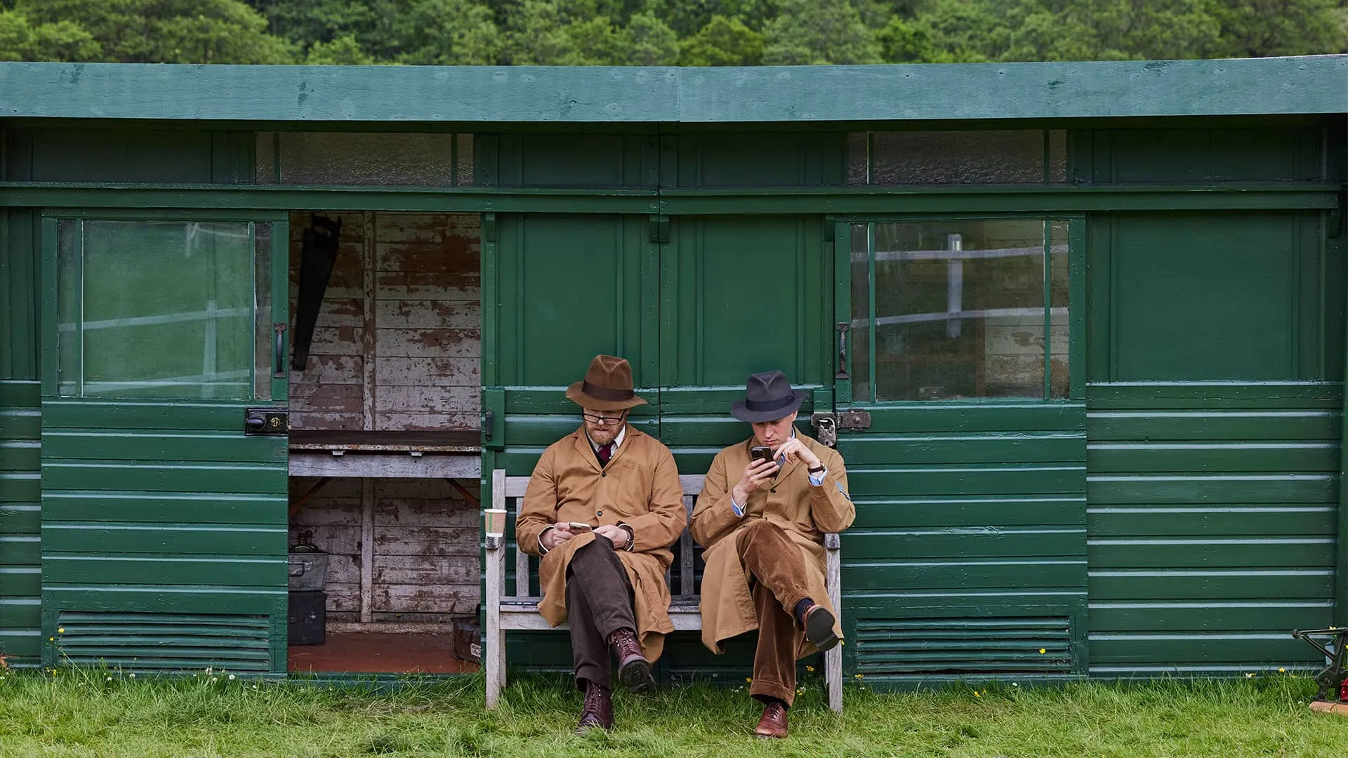 In costume, in a quiet moment, Samuel West and Nicholas Ralph, in costume, sit next to one another in front of a green barn, looking at their phones.