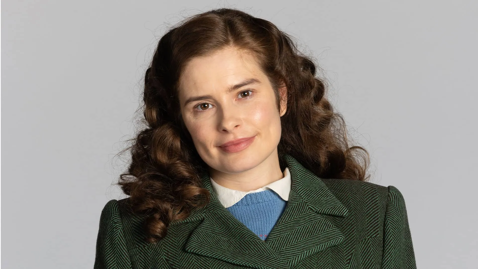 Head and shoulders portrait of Rachel Shenton as Helen Herriot, slightly smiling and looking directly at the camera, wearing a green tweed coat and a blue sweater beneath