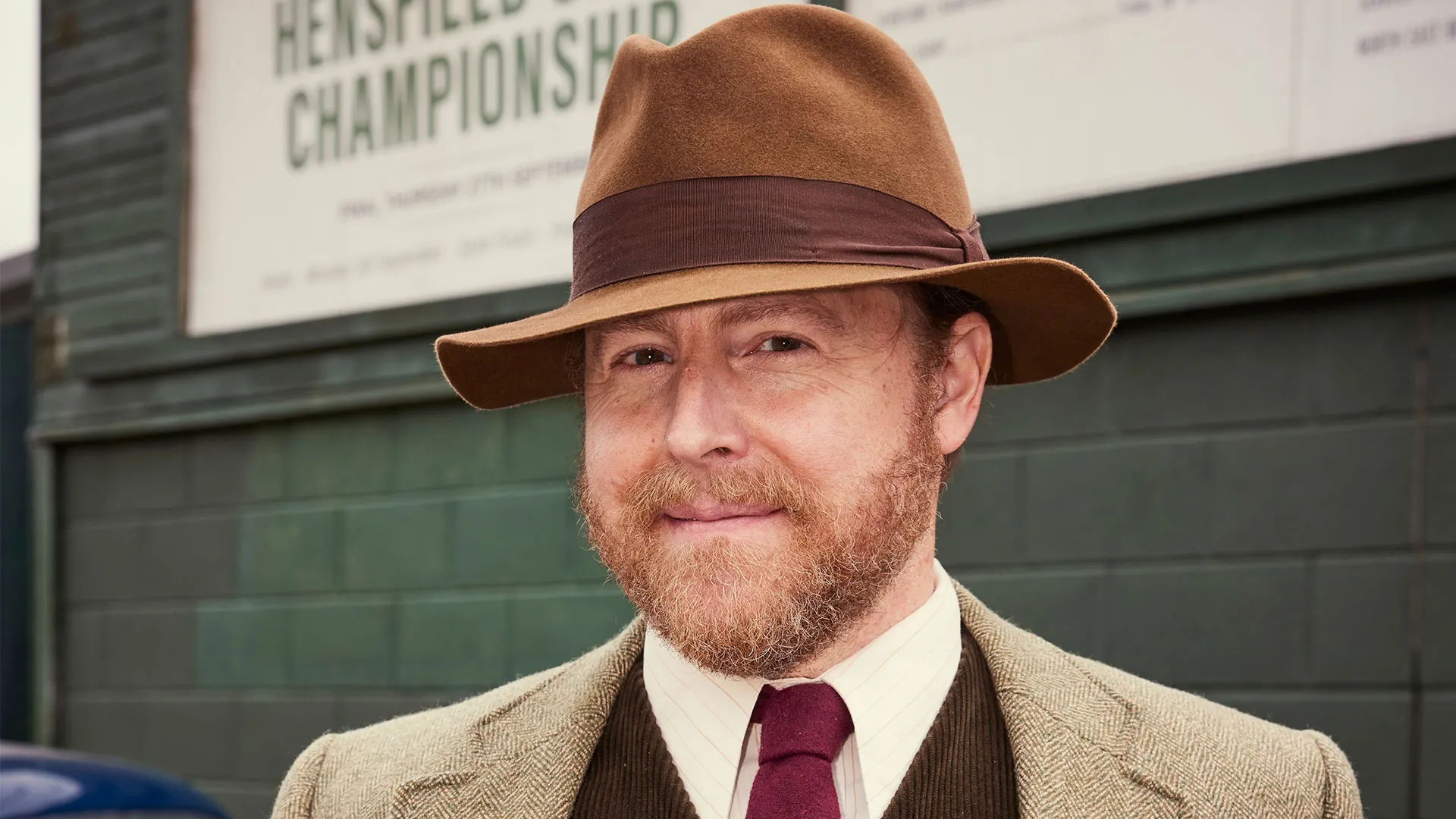 Samuel West as Siegfried Farnon, smiling and wearing a tweed jacket and hat, outdoors in Season 6 of All Creatures Great and Small