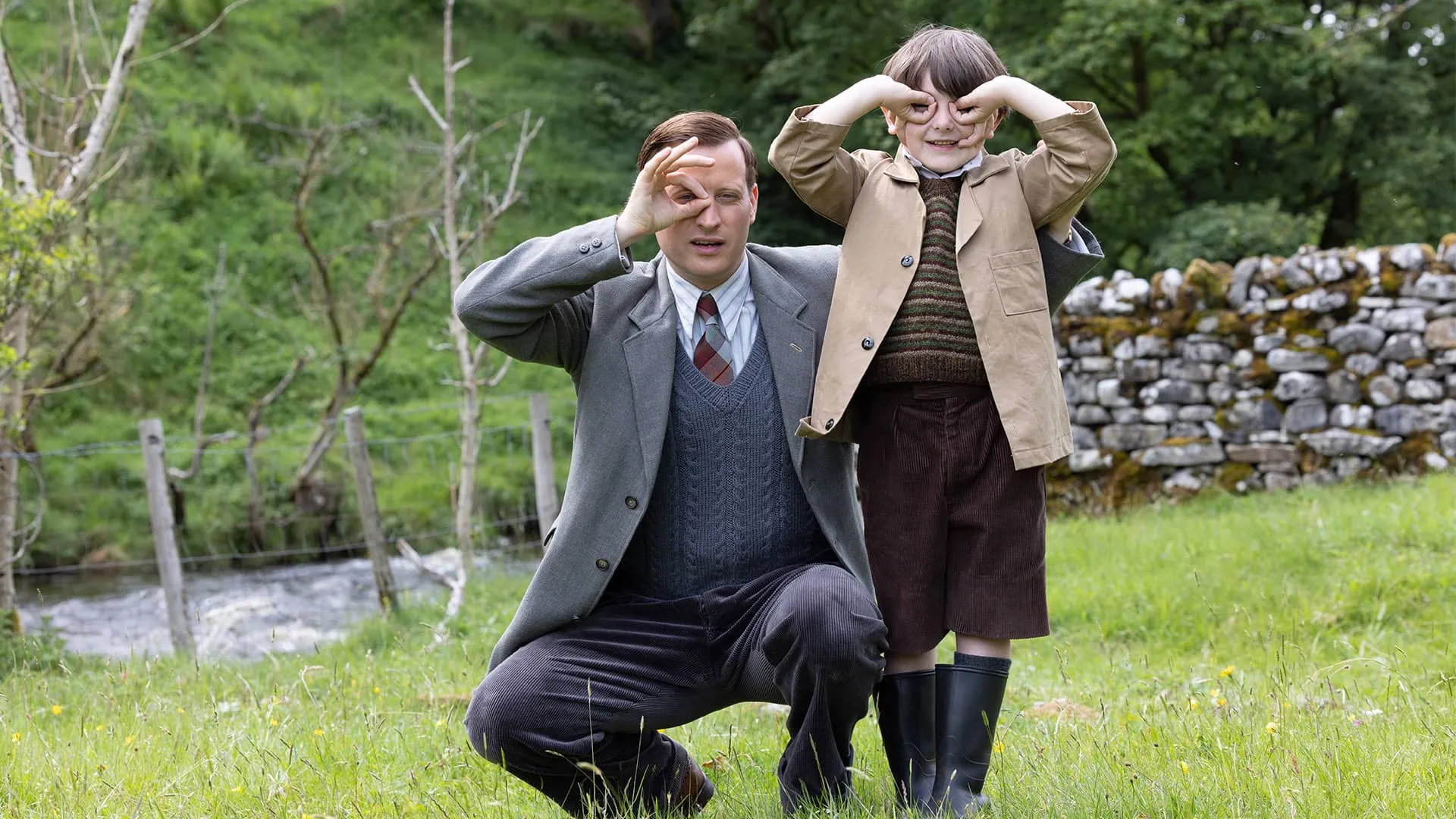 Nicholas Ralph and Thomas Riches side-by-side outdoors. Nicholas uses his thumb and forefinger to make a monacle over his eye and Thomas uses his fingers to make a birdman mask over his eyes