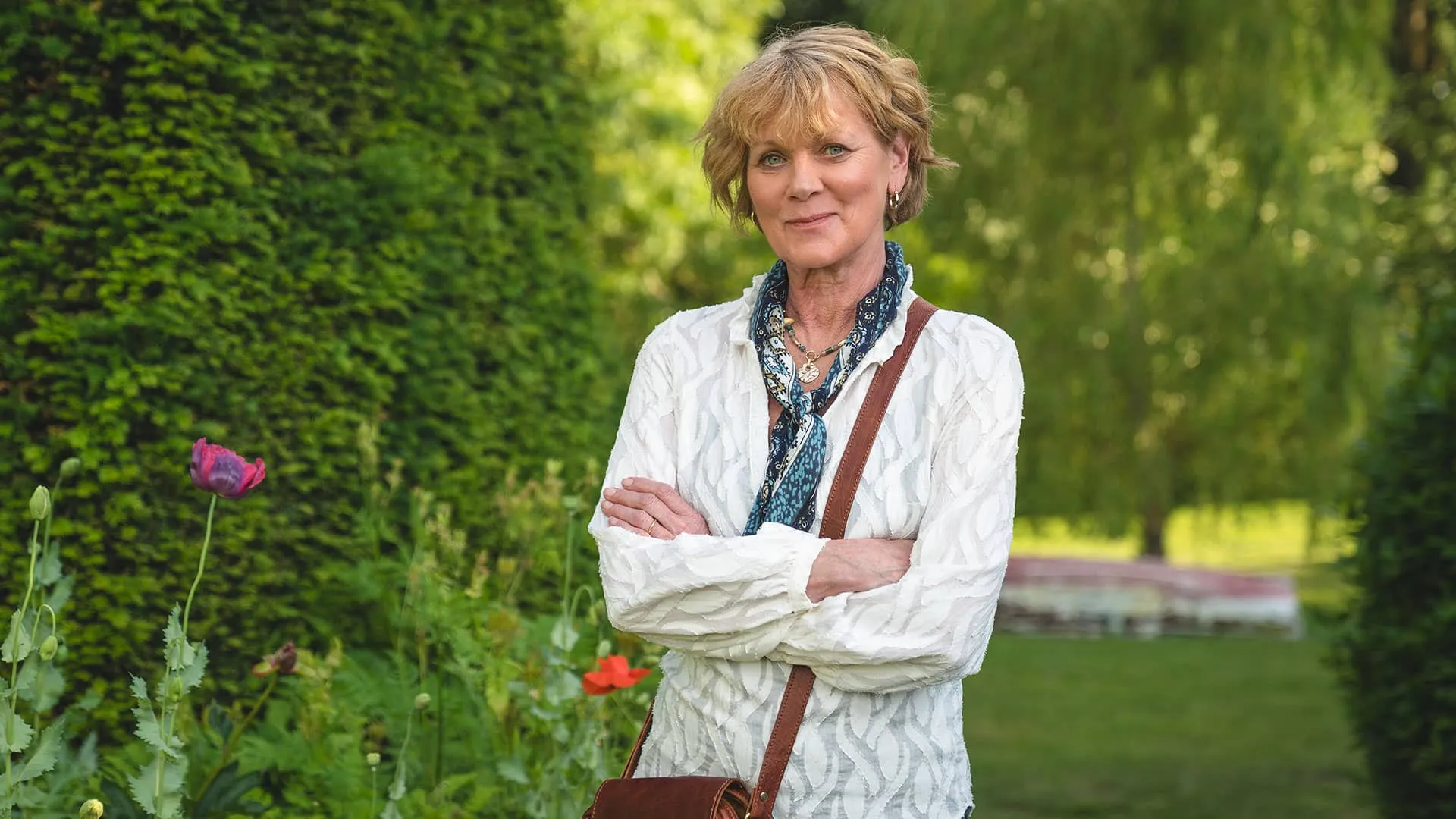 Arms crossed, smiling, Judith Potts (Samantha Bond) poses in sunny, flowery outdoors
