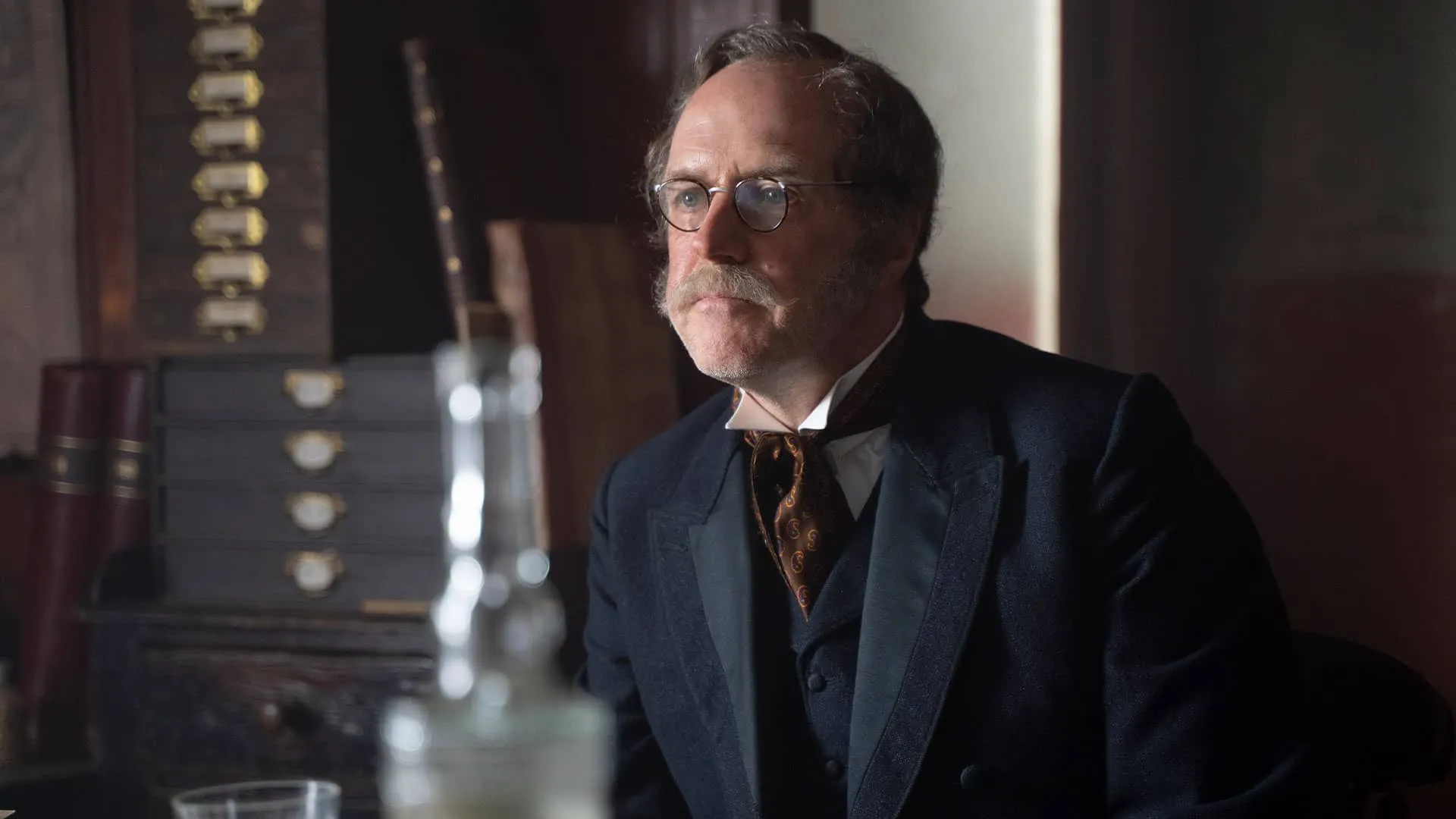 Henry Scarlet (Kevin Doyle), bespectacled in Victorian attire, looks off camera in Miss Scarlet Season 1