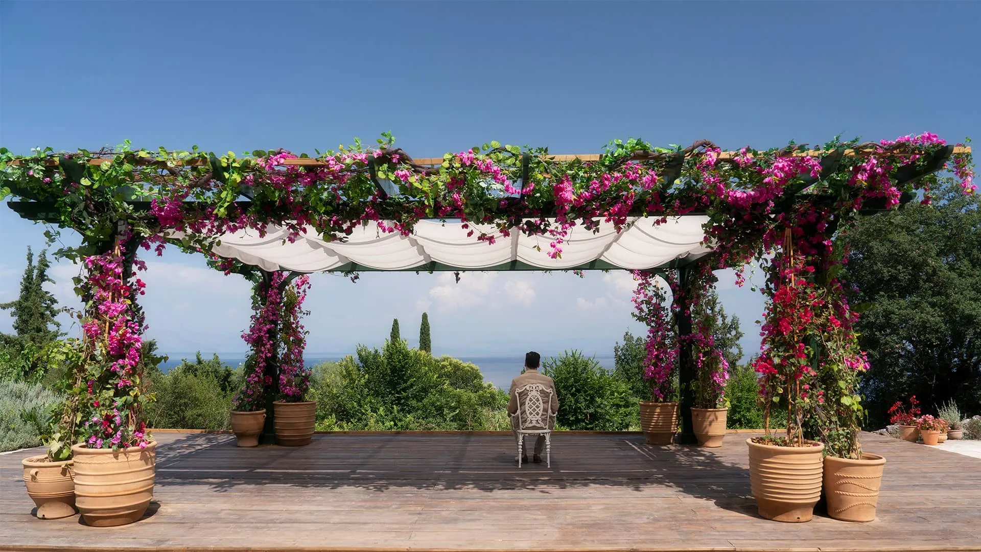Detective Atticus Pund sits alone outdoors under a flowered pergola, gazing at the sea