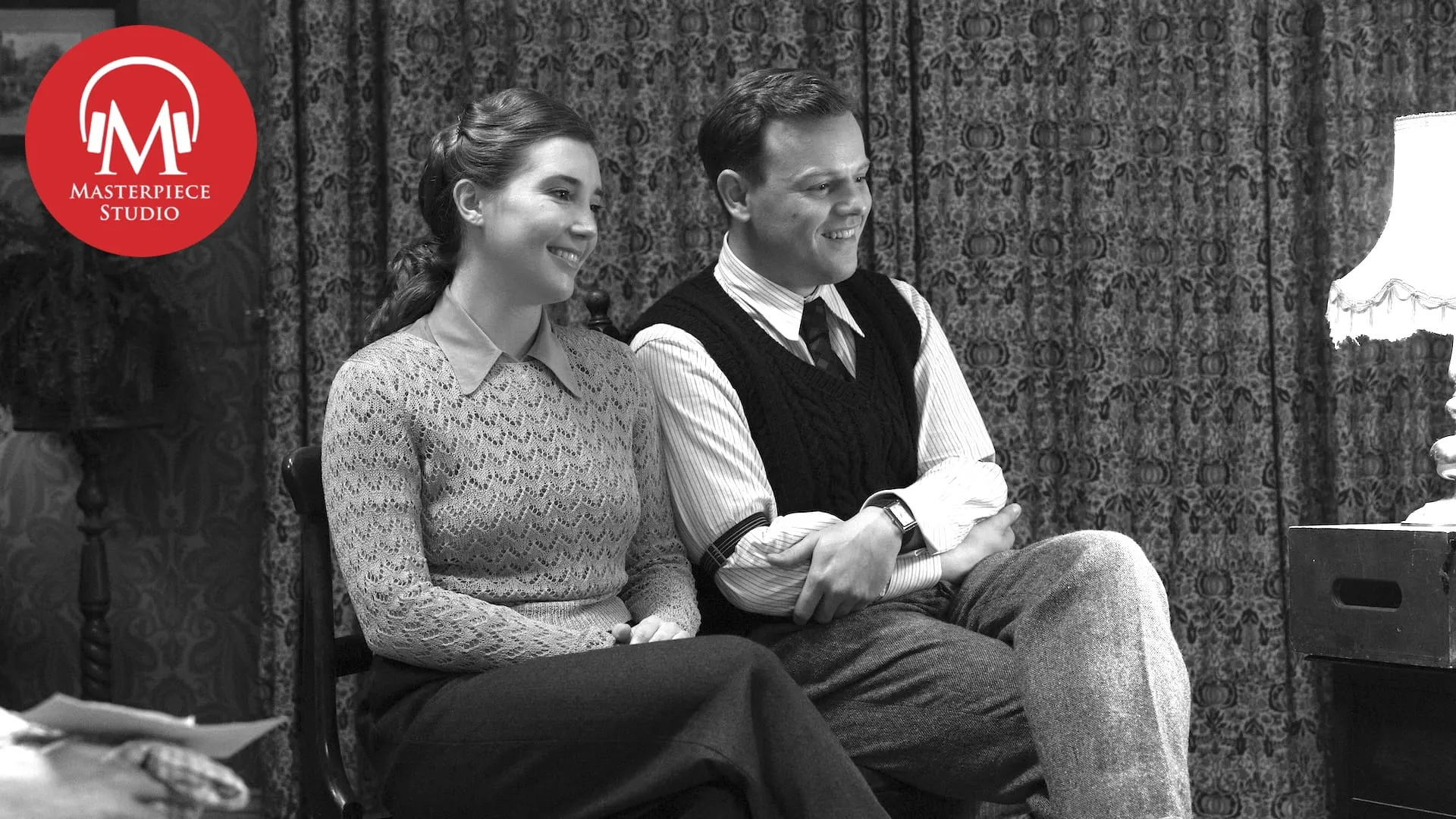 black and white photo of actors callum woodhouse and gaia wise in all creatures great and small season 7, sitting in chairs in a living room. the red masterpiece studio podcast logo is in the top left
