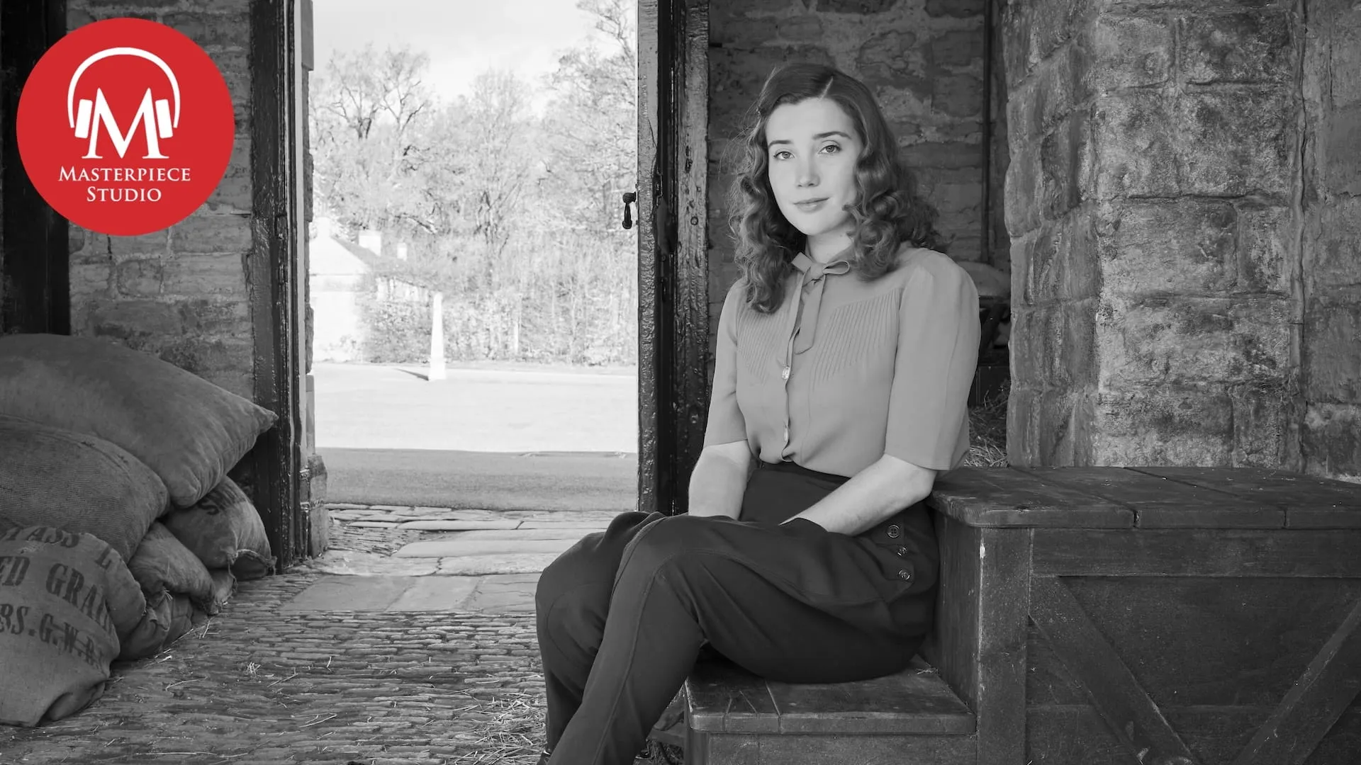 Black and white photo of actor Gaia Wise sitting down in a barn looking at the camera with the red MASTERPIECE Studio logo in the top left