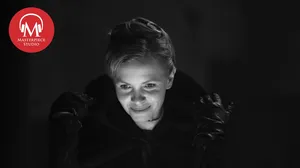 This is a black and white photo of actor Kate Phillips from Season6, Episode 1 of Miss Scarlet. She is taking off her hood and her face is illuminated. There is a red MASTERPIECE Studio logo top left