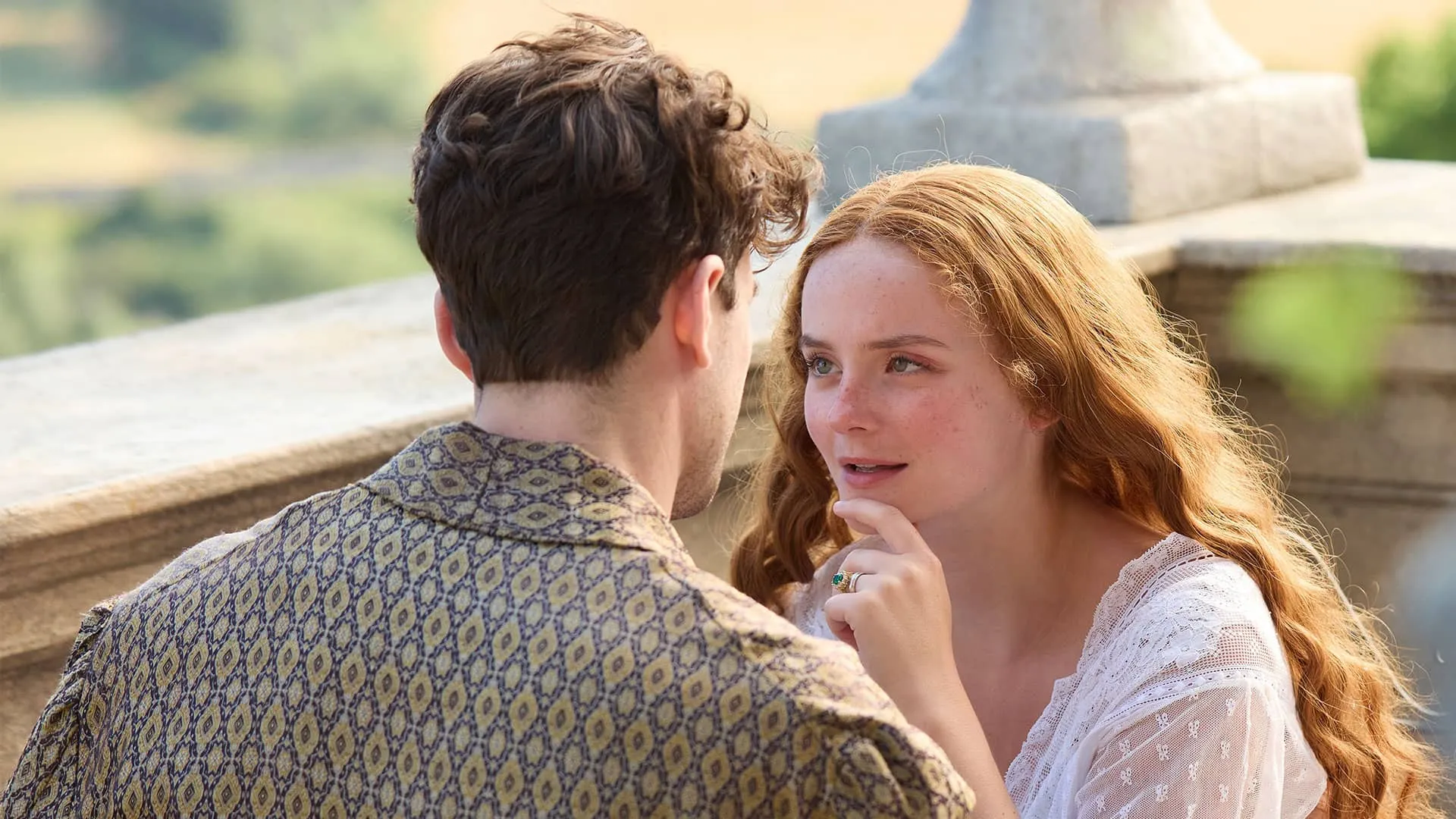 Soames (with back to the camera) and Irene sit together on a balcony, both in robes, in an intimate moment. With her long red hair down, smiling slightly, she looks lovingly into his eyes.