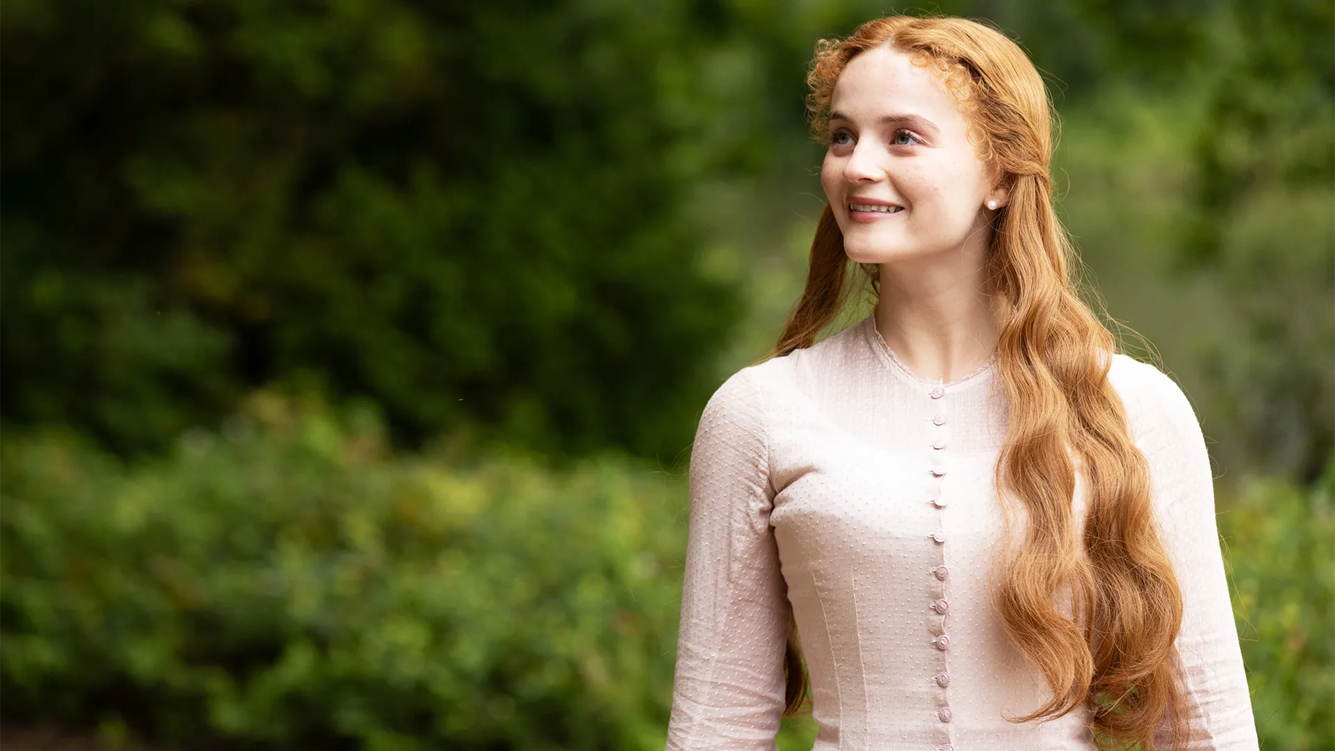 British actor Millie Gibson as Irene Heron in a pink dress against a garden backdrop in a scene from The Forsytes Season 1 on MASTERPIECE on PBS.