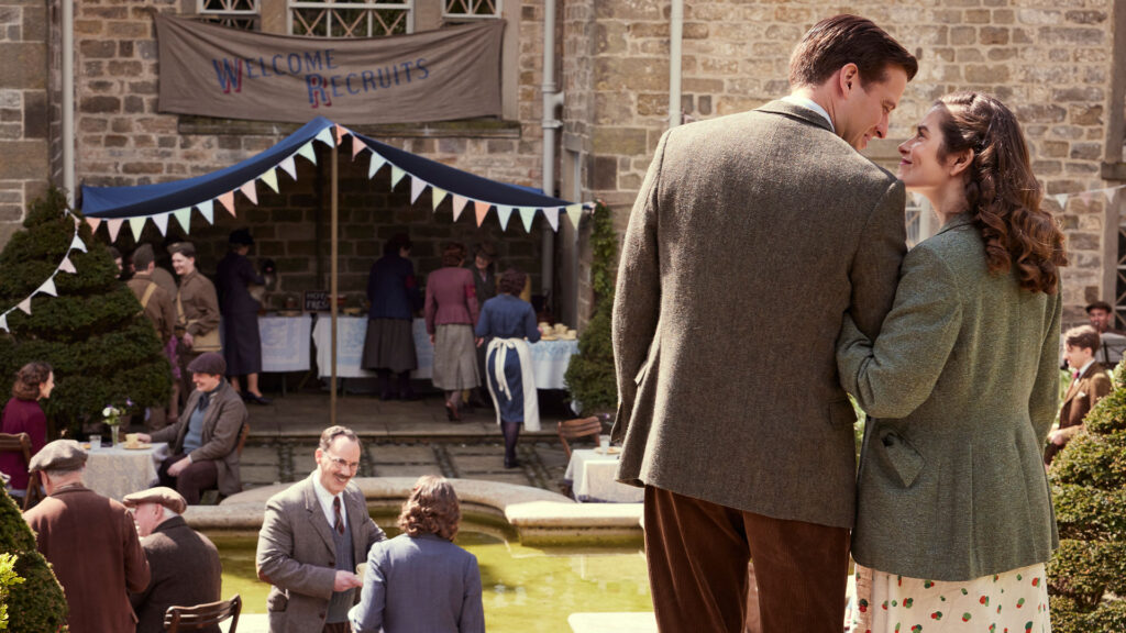 James and Helen look lovingly at each other standing outside of an outdoor party with bunting and a sign reading 