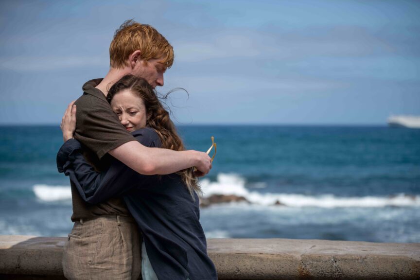 Domhnall Gleeson as Jack and Andrea Riseborough as Alice hugging by the ocean in a scene from Alice & Jack, a miniseries from MASTERPIECE on PBS.
