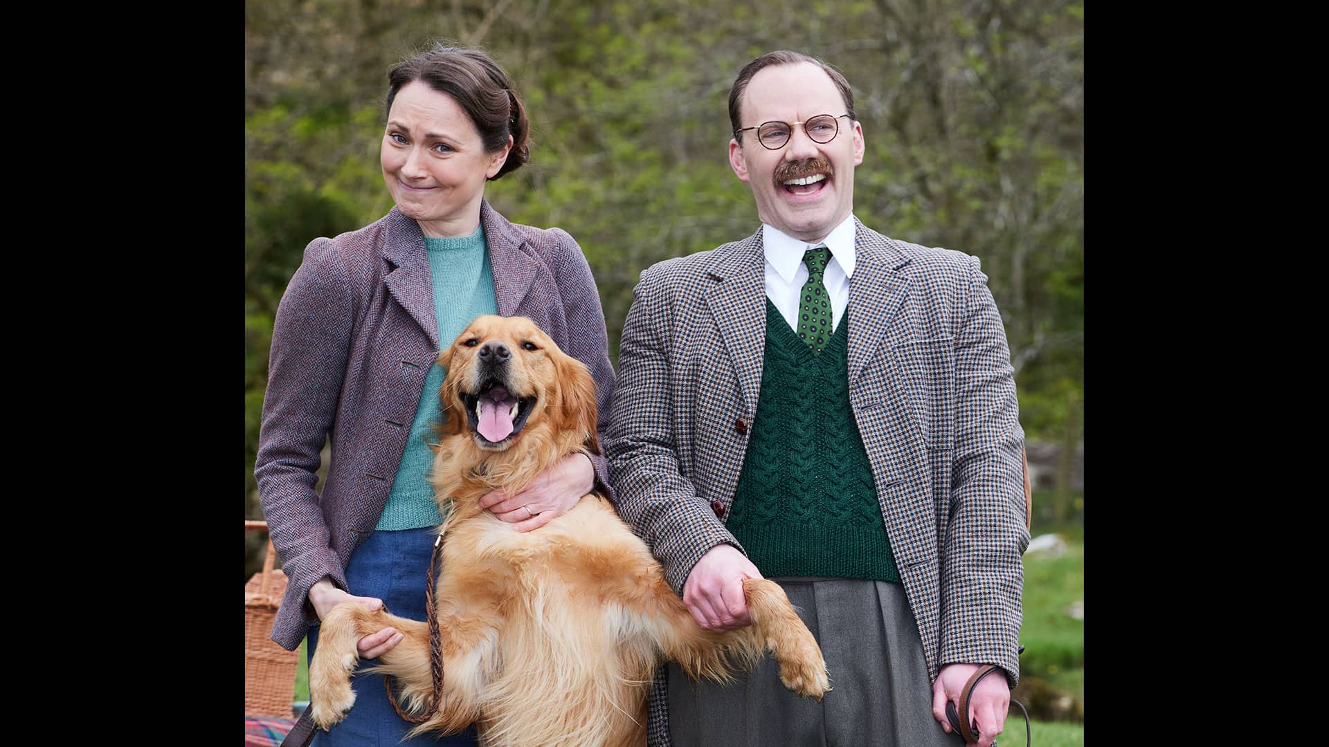 actress Anna Madeley (Mrs. Hall) and Will Thorpe (Gerald) tand outdoors with golden retriever Ernie (Jess) on his hind legs between them, looking happy with his tongue out. Will laughs and Anna smiles in a knowing way.