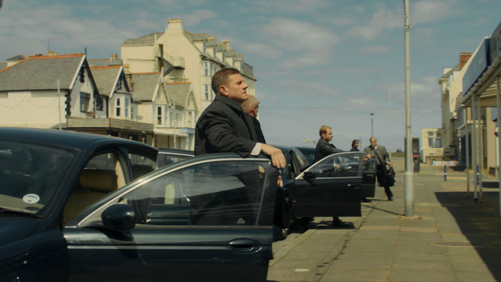 Scene from the PBS miniseries Mr Bates vs The Post Office with auditors exiting their cars as they call on Subpostmaster Alan Bates.