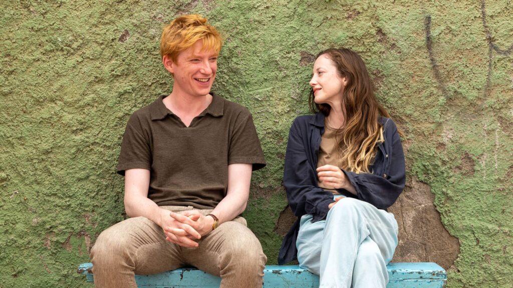 Jack and Alice seated on a blue bench in front of a green wall in a lighthearted moment