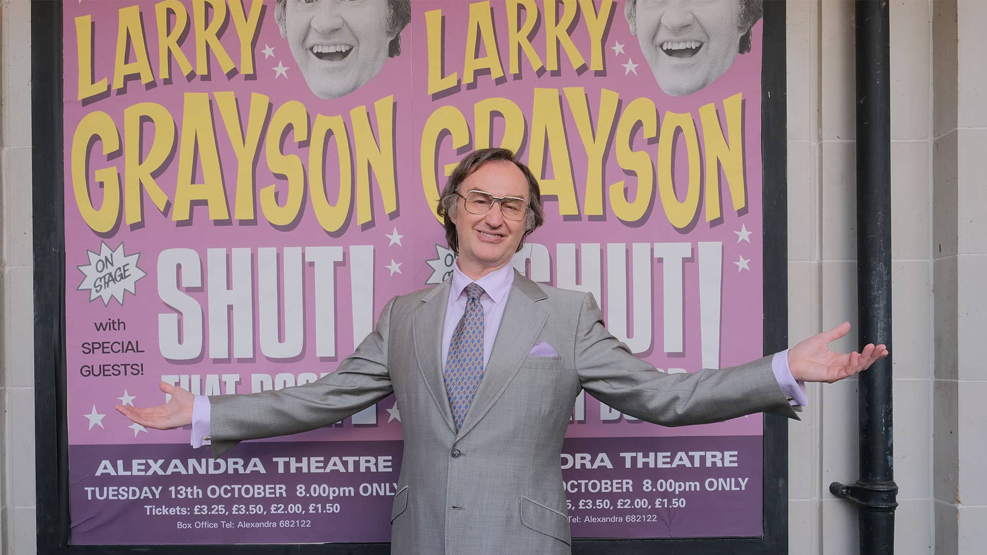 Mark Gatiss as Larry Grayson, smiling and posing in front of a giant pink and yellow poster promoting his show