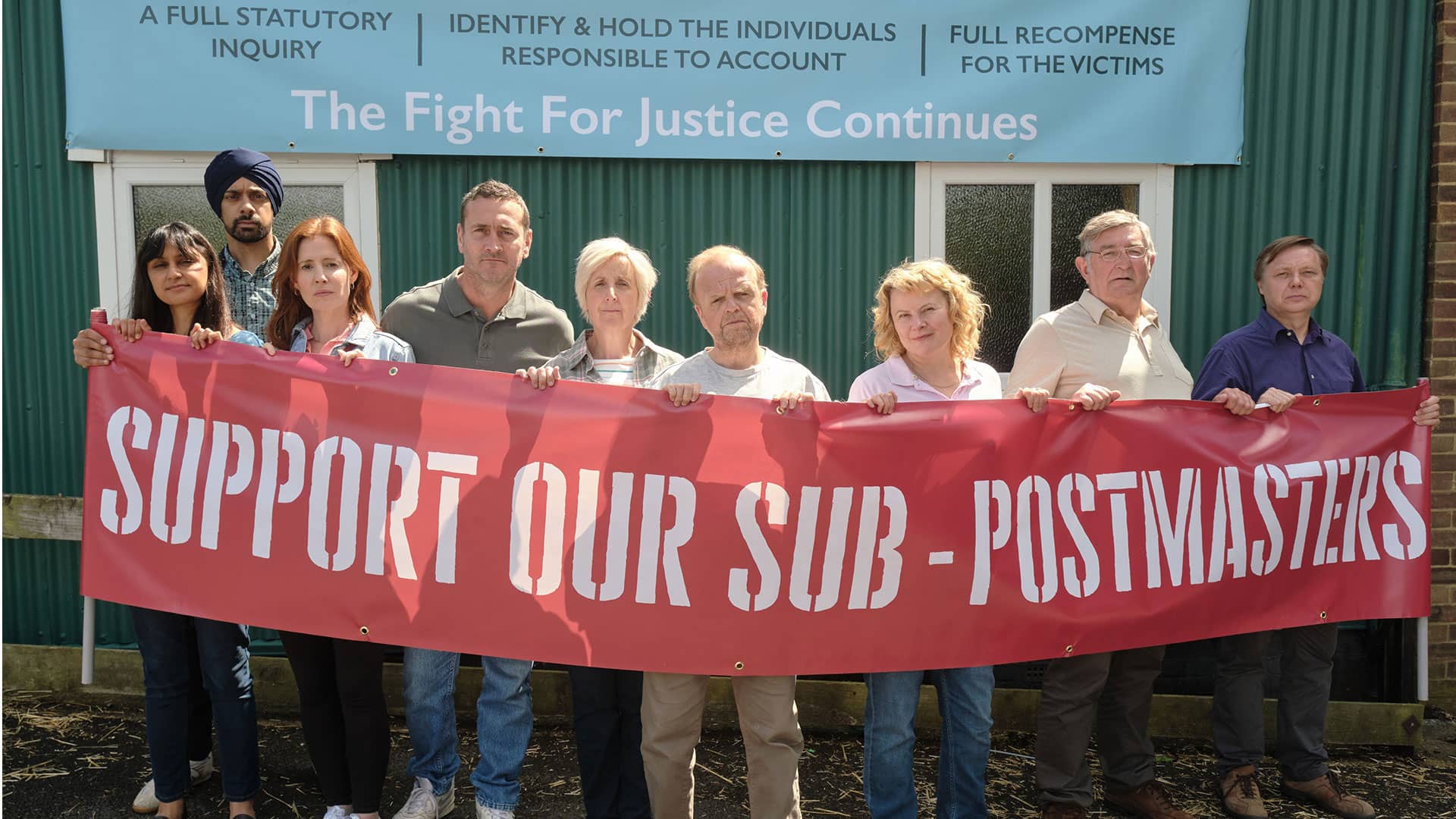 Alan Bates and other Subpostmasters and spouses stand outdoors, defiantly holding a banner saying "Support our Subpostmasters"