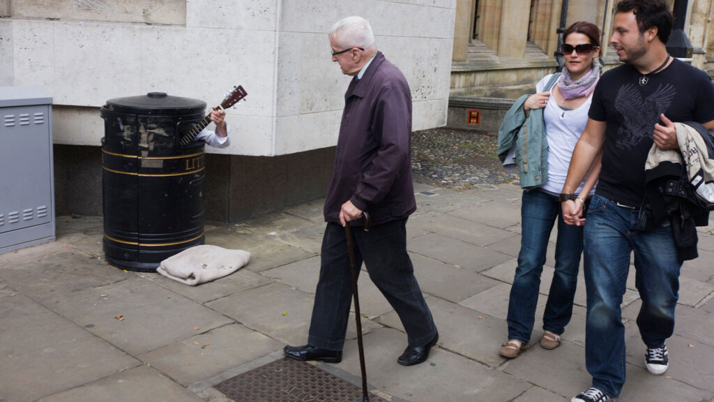Color photo of a street busker hiding in a litter bin to earn extra cash as passers-by hear his music on a Cambridge pavement.