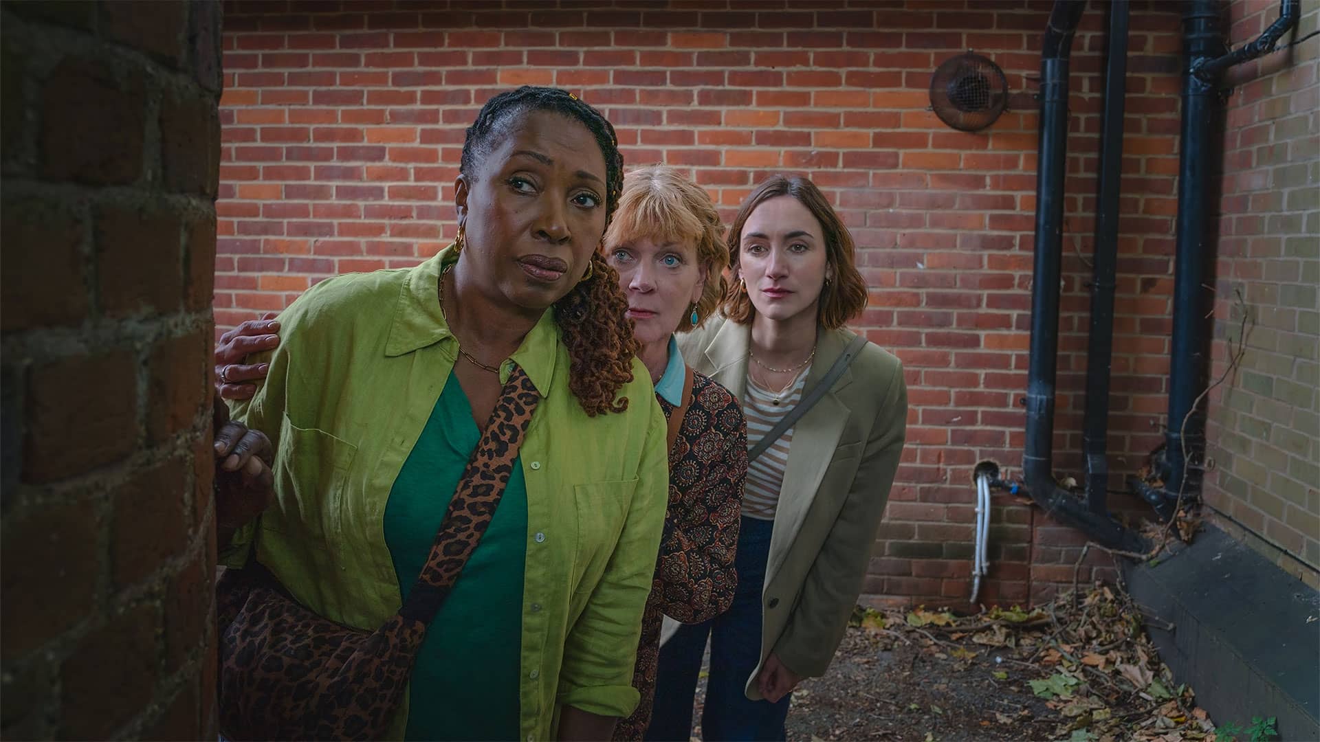 Suzie Harris, Judith Potts, and Becks Starling peek tentatively around the corner in a brick alley