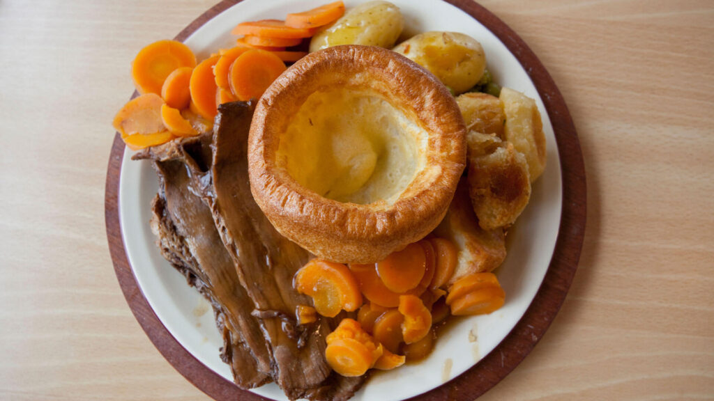 An English roast dinner on a plate with orange carrots, roast beef with gravy, boiled potatotes and a golden brown Yorkshire pudding in the middle.