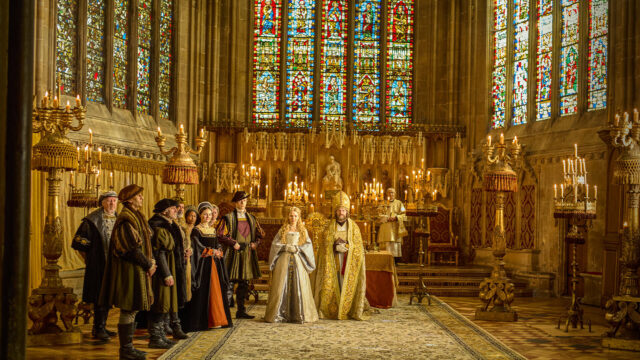 Scene from Wolf Hall: The Mirror and the Light with Kate Phillips as Jane Seymour awaiting Damian Lewis as Henry VIII for their wedding ceremony. They are in a chapel with soaring ceilings, candelabra, and stainedglass windows. The historical drama airs on MASTERPIECE on PBS.