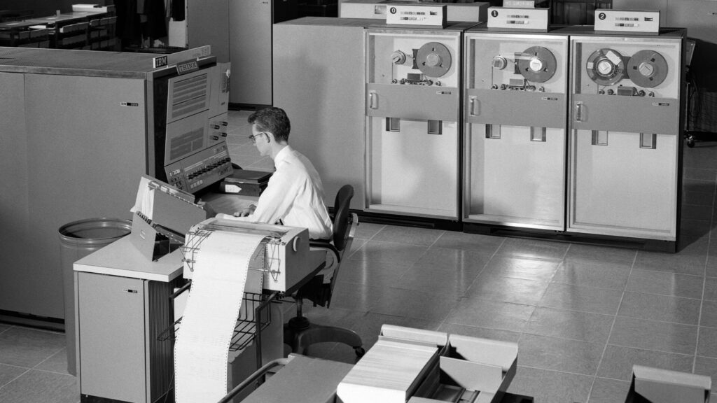 Black and white photo of man Man programming large mainframe computer surrounded by data tape drives in early 1960s.