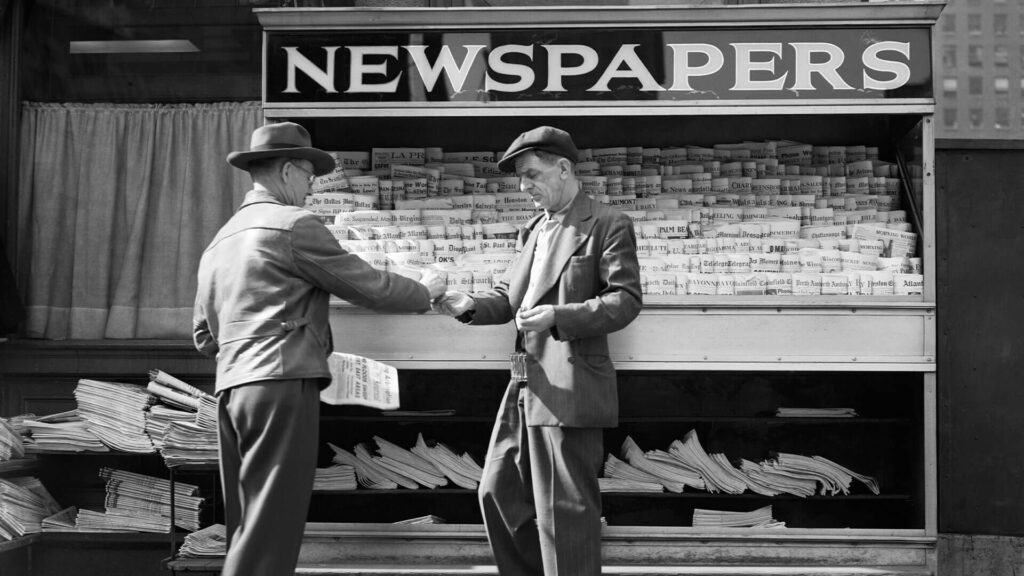 Black and white photo from the 1940s of a man buying a newspaper from a sidewalk vendor in New York City.