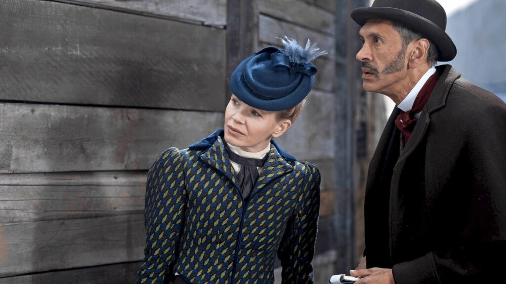 Color image of a scene from Miss Scarlet Season 6 on MASTERPIECE on PBS. Actor Kate Phillips as Eliza Scarlet stands next to Paul Bazely as her investigative office's bookkeeper, Clarence.