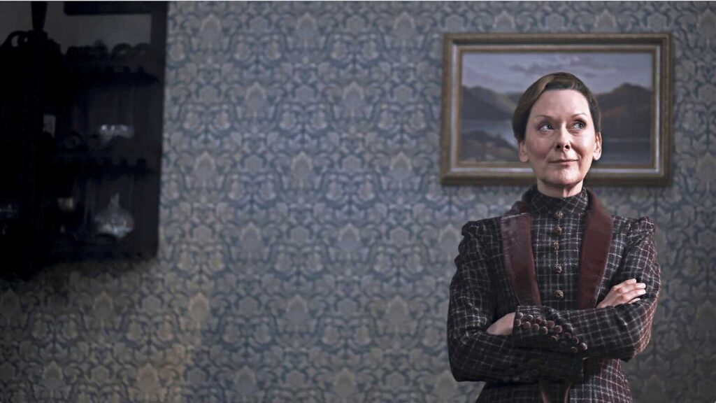 Color image of a scene from Miss Scarlet Season 6 on MASTERPIECE on PBS. Actor Cathy Belton stands with arms crossed in her role as housekeeper, Ivy.